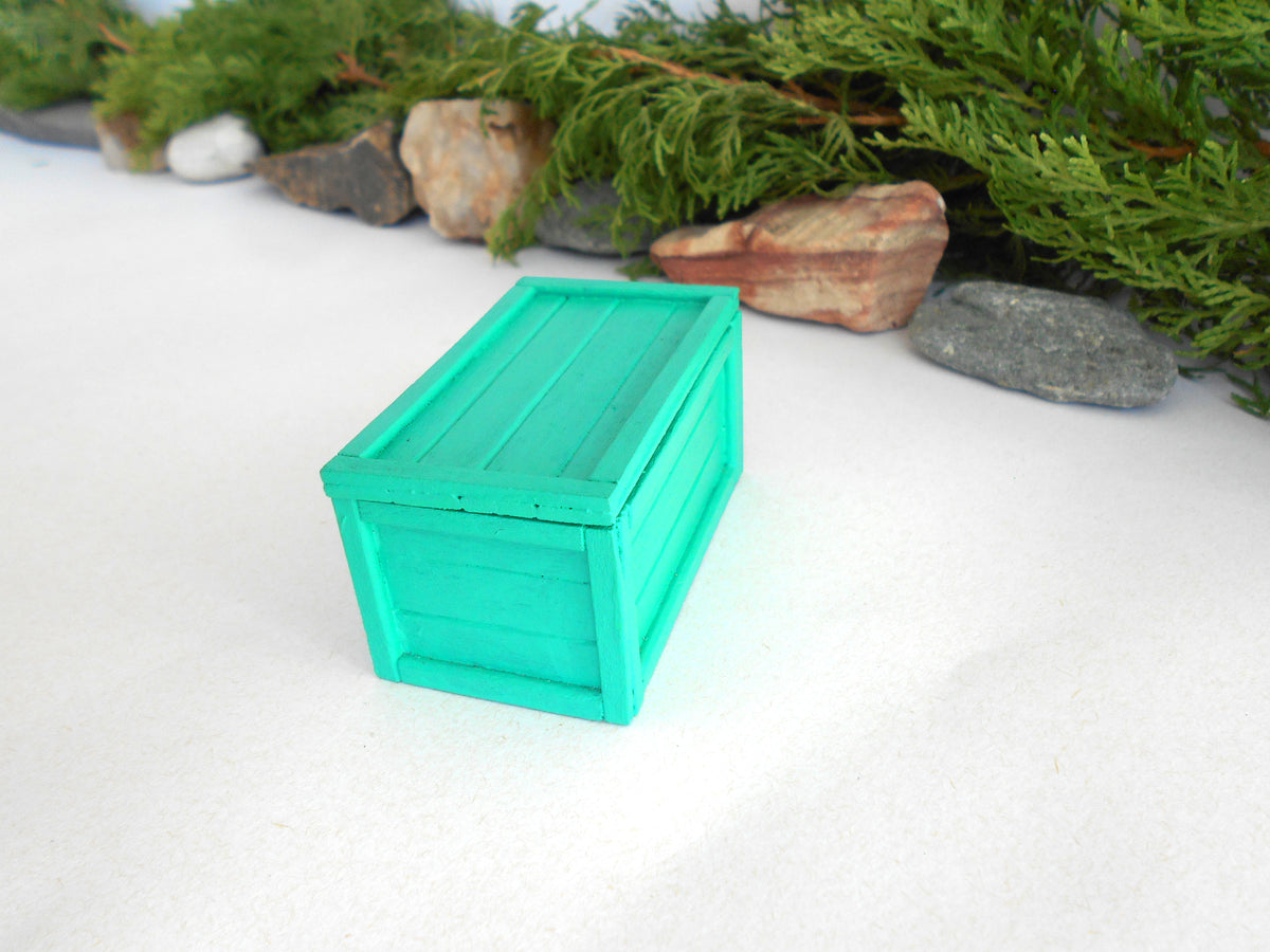 A miniature bright green wooden storage box with a slatted front and lid, set on a white surface with decorative greenery and stones in the background.