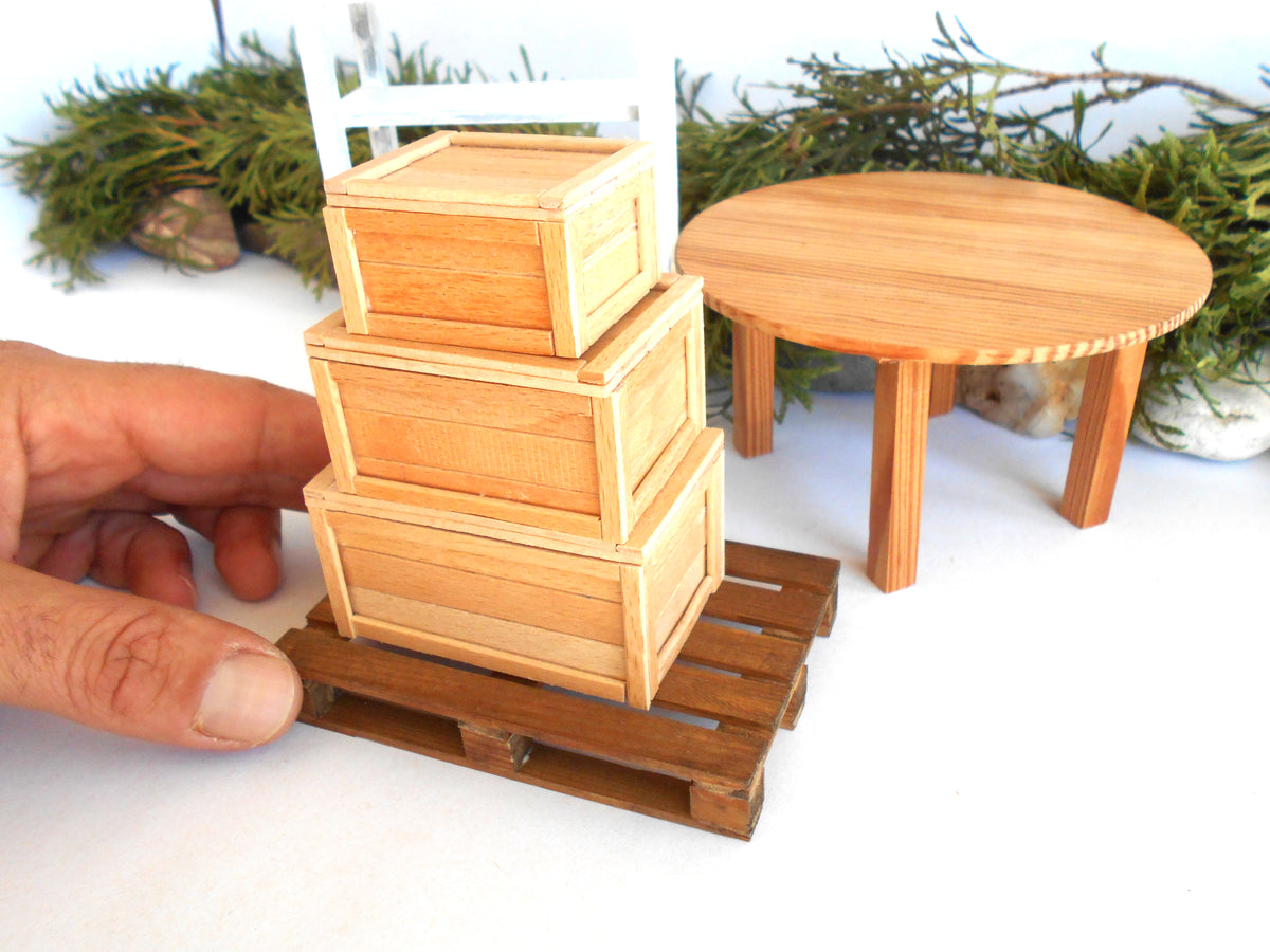 A miniature wooden crate set with three stacked boxes on a small wooden pallet, held by a hand for scale, with a miniature round table and green garlands in the background.