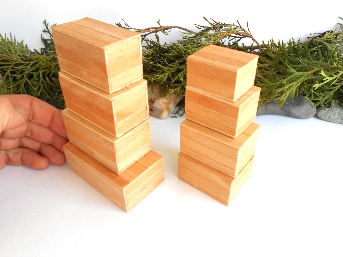 Two stacks of light natural wood rectangular boxes , with a hand holding the left stack to show size and scale, set against a white background with green pine branches.
