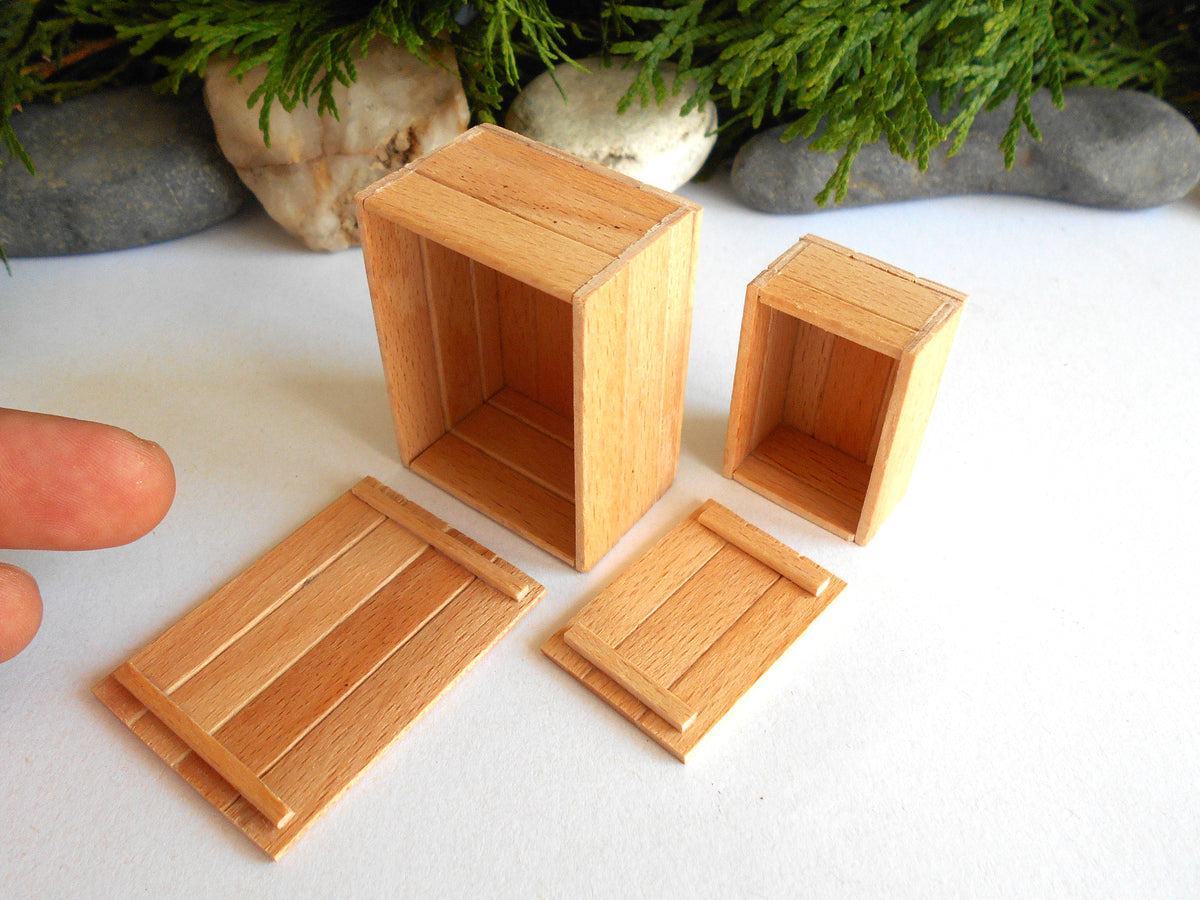 Four miniature light brown wooden boxes and lids, likely for dollhouse or scale model use, displayed on a white surface with decorative greenery and stones in the background.