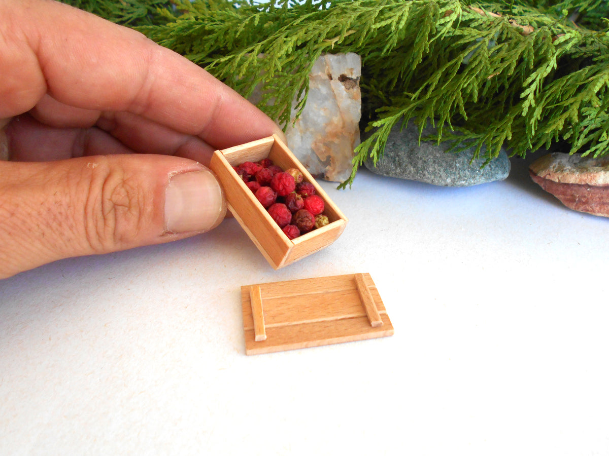 A miniature wooden box filled with small red berries, held by a hand, with its lid placed below on a white surface. Green foliage and small rocks decorate the background.
