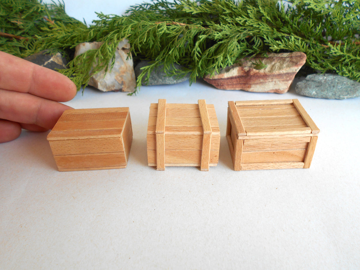 Three miniature light wood crates of different designs displayed on a white surface, with a hand pointing to the leftmost crate and green foliage in the background.