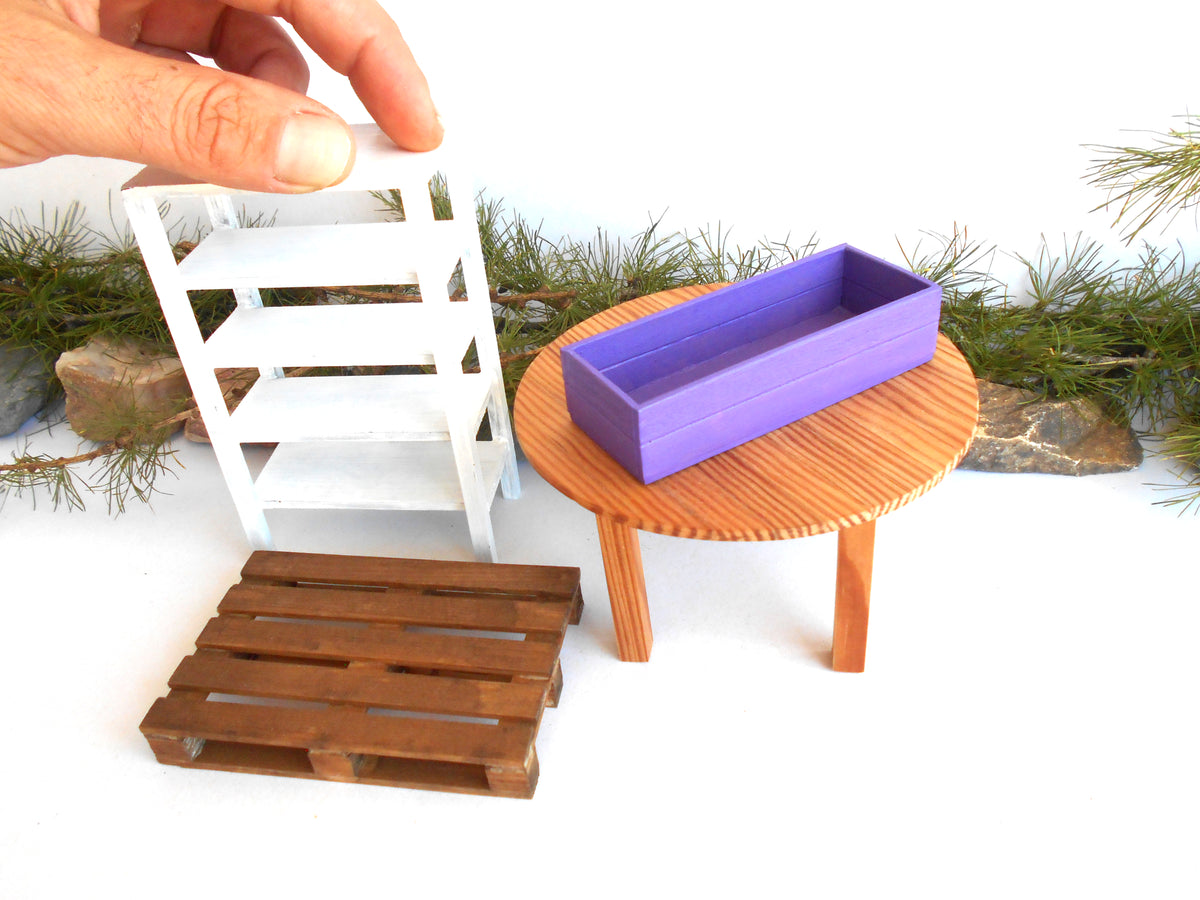 A miniature dollhouse furniture set featuring a white three-tier shelf, a round wooden table with a purple rectangular planter, and a brown wooden pallet, with a hand holding the shelf on a white background.