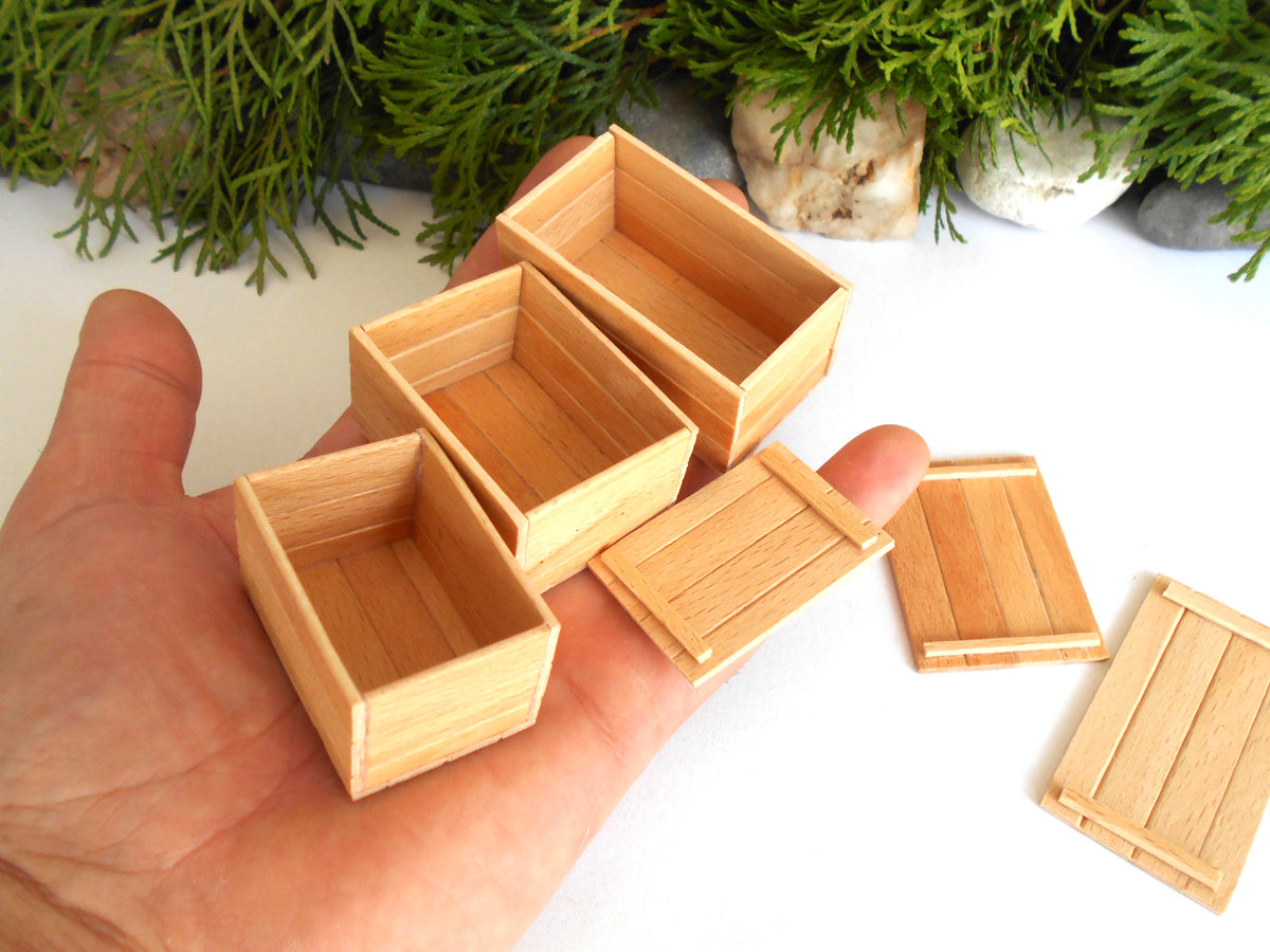 A hand holds several miniature light wood boxes and lids, with more boxes placed on a white surface nearby and green foliage in the background.