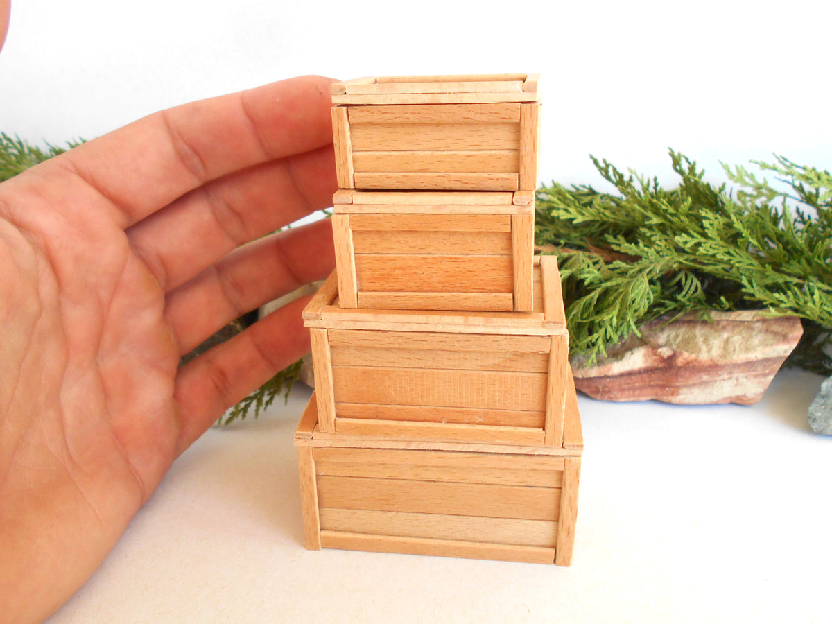 A hand holding a stack of three miniature light wood crates, shown against a white background with small green decorative plants in the background.