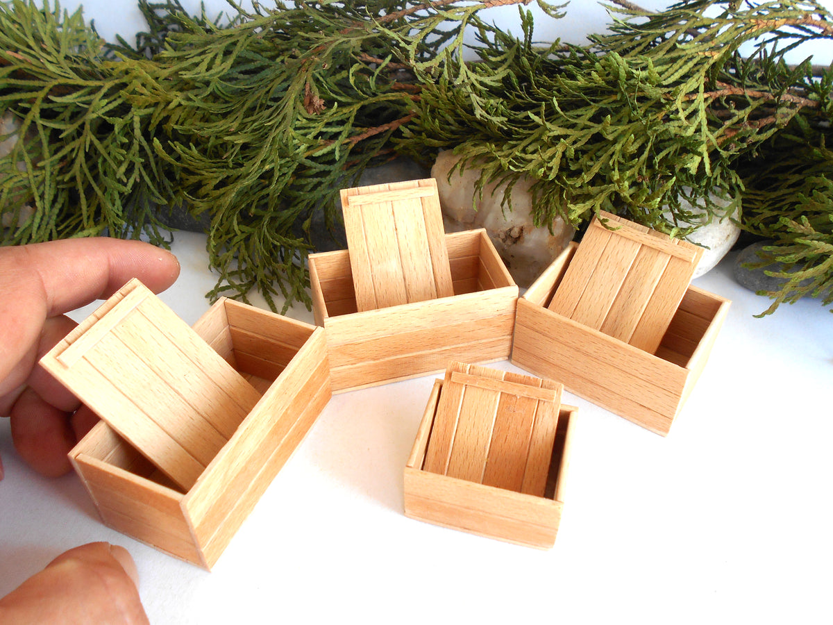 A set of miniature light wood boxes with sliding lids, shown with a hand adjusting one of the boxes. The boxes are arranged on a white surface with green foliage in the background.