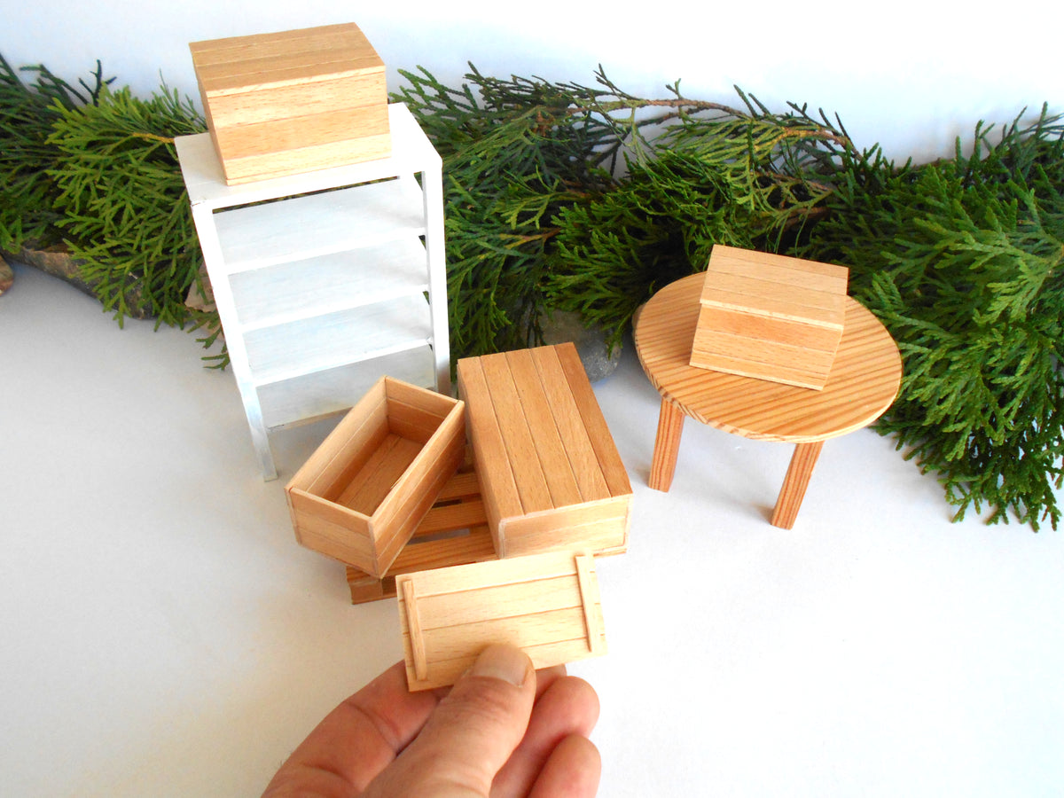 A hand holds a small wooden miniature crate in the foreground, with several other matching wooden miniature furniture pieces arranged behind it on a white surface, including a cabinet, round table, and boxes. Green foliage decor is visible in the background.