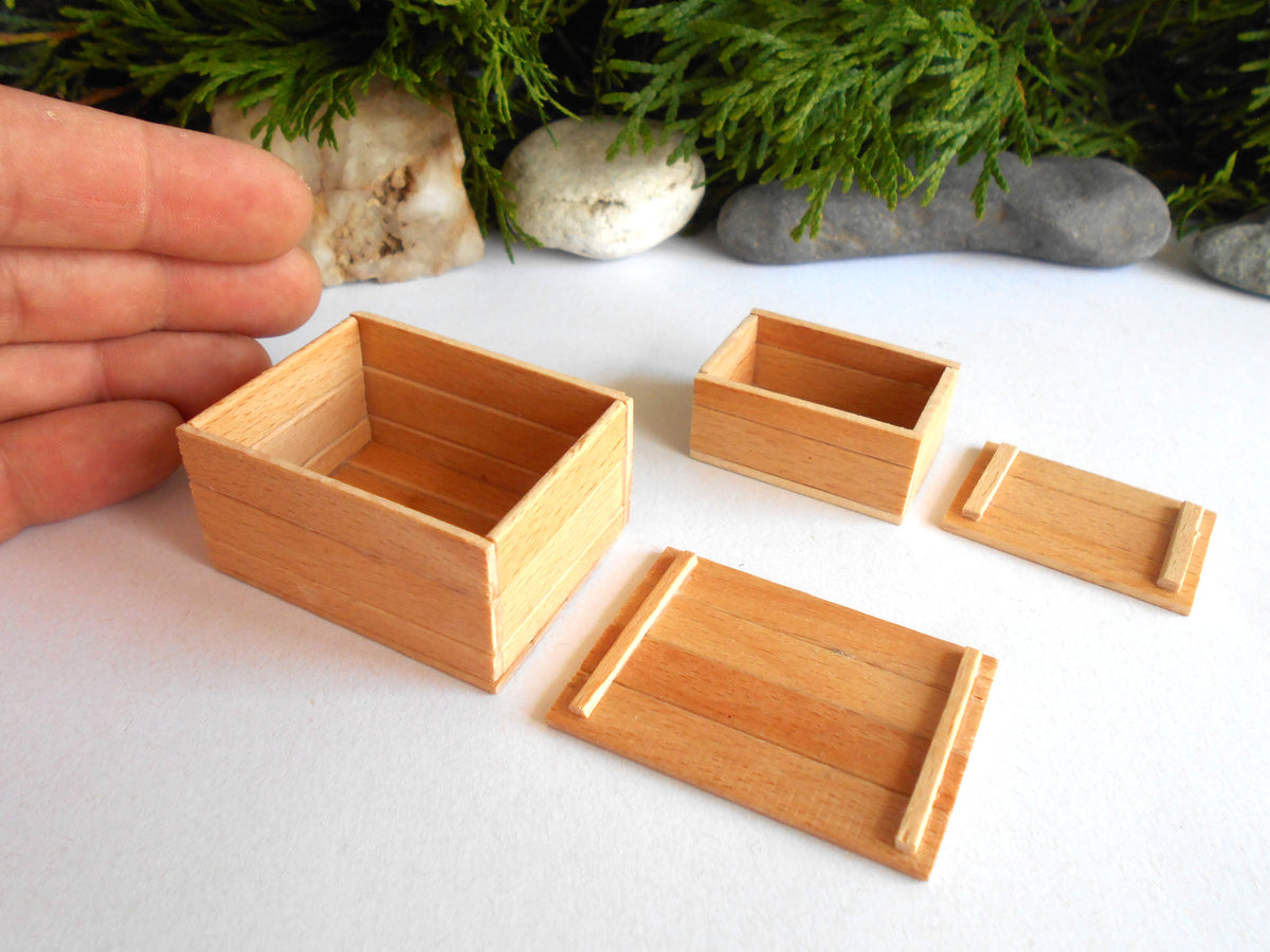 Four miniature light wood boxes and lids arranged on a white surface, with a person's hand visible on the left for scale and greenery and stones in the background.
