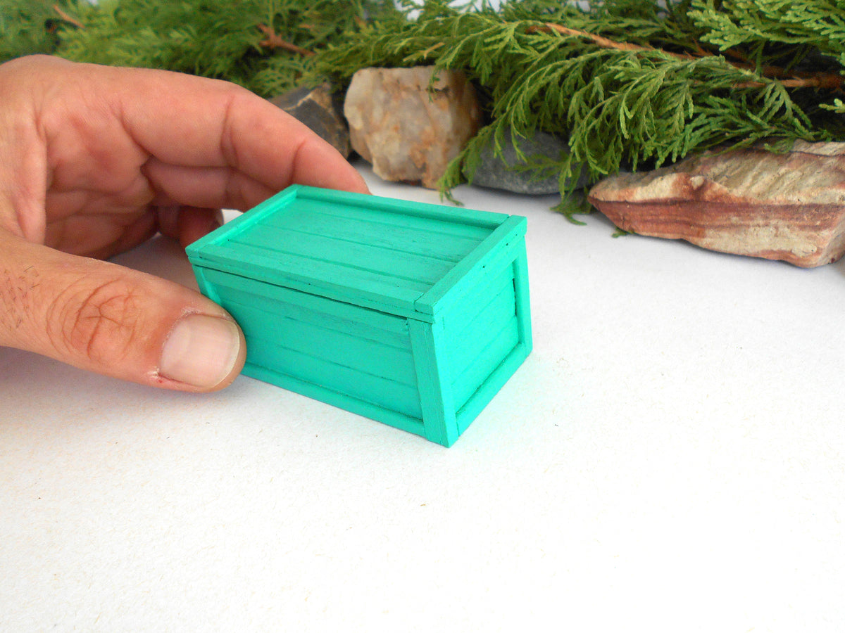 A hand holding a small, bright turquoise-green wooden crate or box on a white surface with green foliage and rocks in the background.