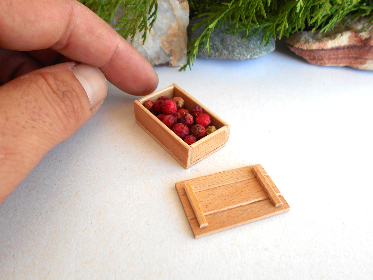 A miniature wooden storage box filled with small red berries, shown next to its lid on a white surface, with a human hand pointing at it for scale.