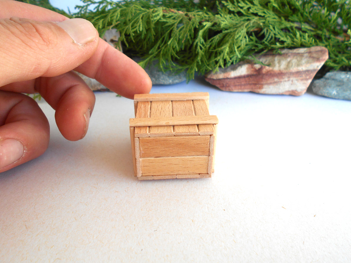 A miniature light wood crate or box shown on a white surface, with a person's fingers pointing at it for scale and green foliage in the blurred background.