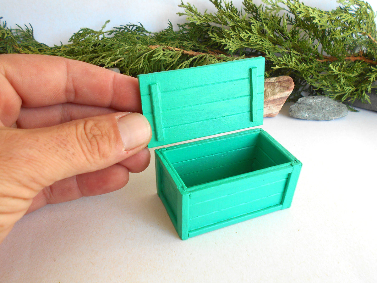 A miniature green wooden storage box with its lid open, held by a hand to show scale, with green foliage in the background.