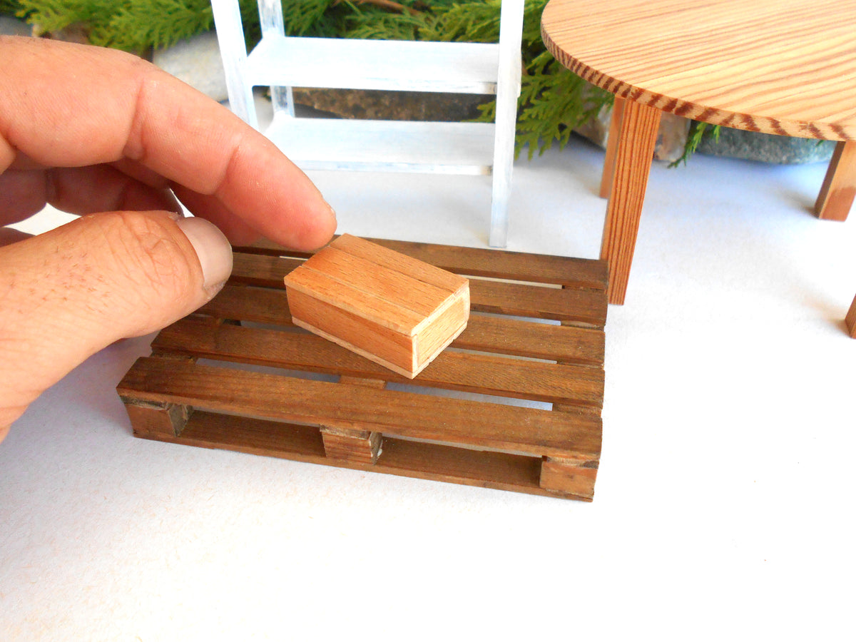 A miniature wooden pallet scale model with a small hand resting on top, shown in a close-up shot with a white surface and miniature furniture in the background.