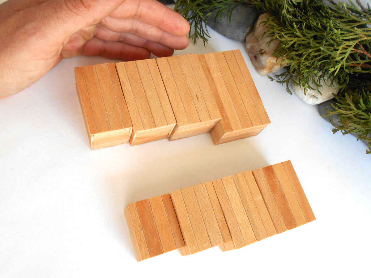 A hand is placing light natural wood boxes onto a white surface, with two sets of rectangular wooden blocks and decorative greenery in the background.
