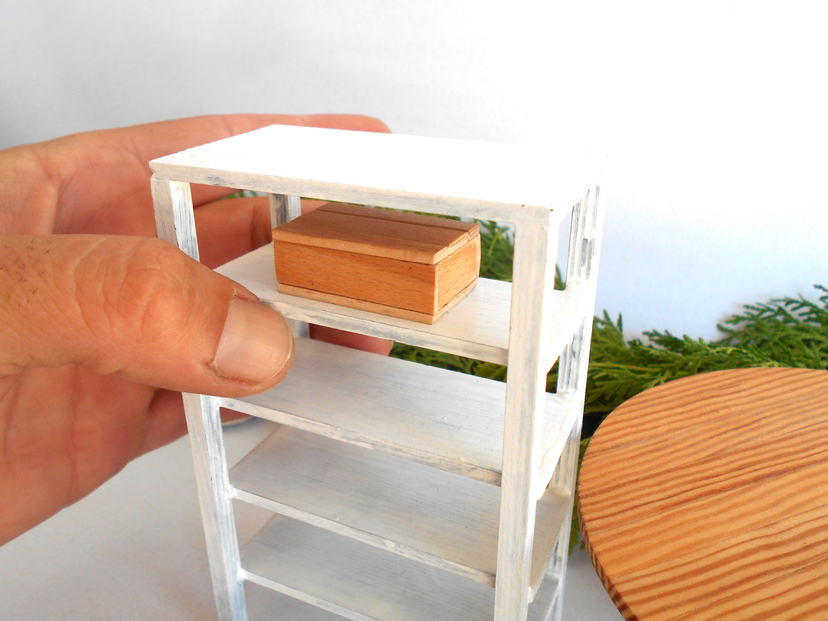 A miniature white shelving unit with a small wooden box on one of its shelves, held by a person's hand to show scale, with a round wooden table in the background.