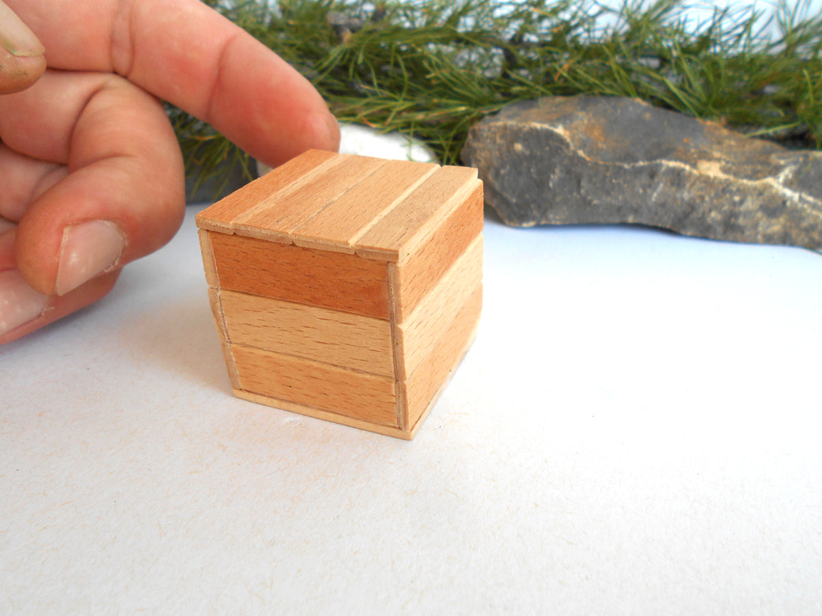A small handcrafted wooden box with a lid, made from light-colored wood with horizontal grain patterns, shown with a person's hand pointing to it on a white surface.