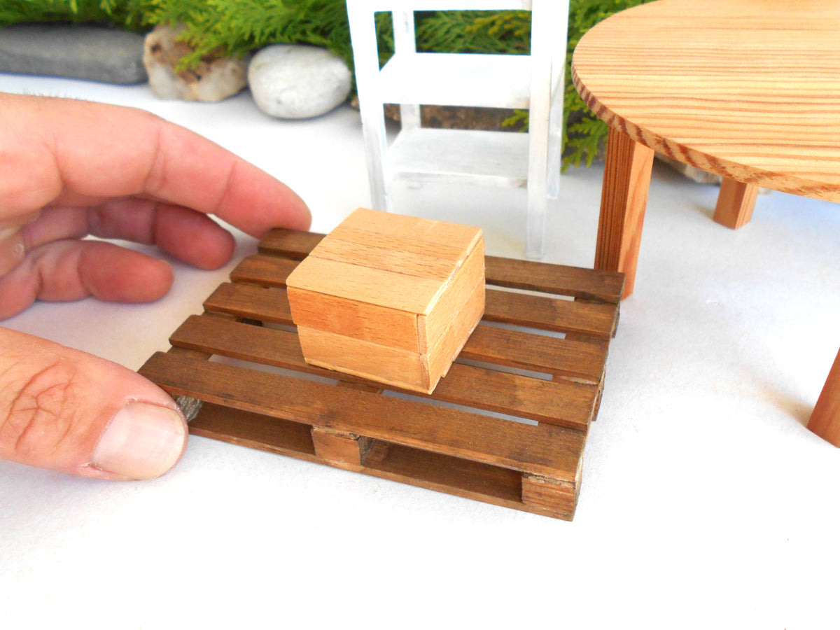 A miniature wooden pallet model with a small wooden box on top, held by a human hand to show scale, with a miniature white chair and round wooden table in the background.