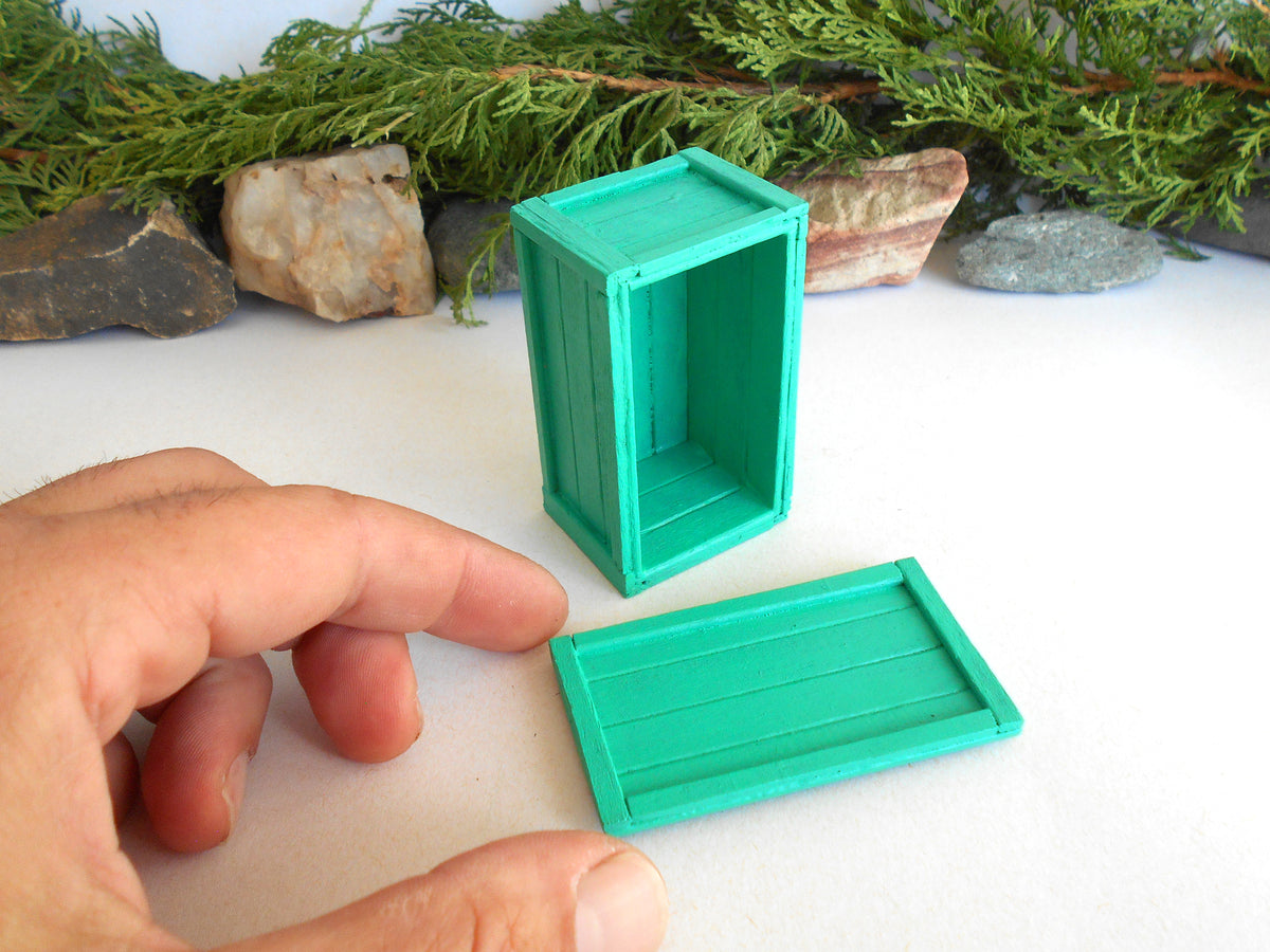 A miniature green plastic crate or box set with a separate lid, shown on a white surface with a hand pointing to it for scale, and greenery and rocks in the background.