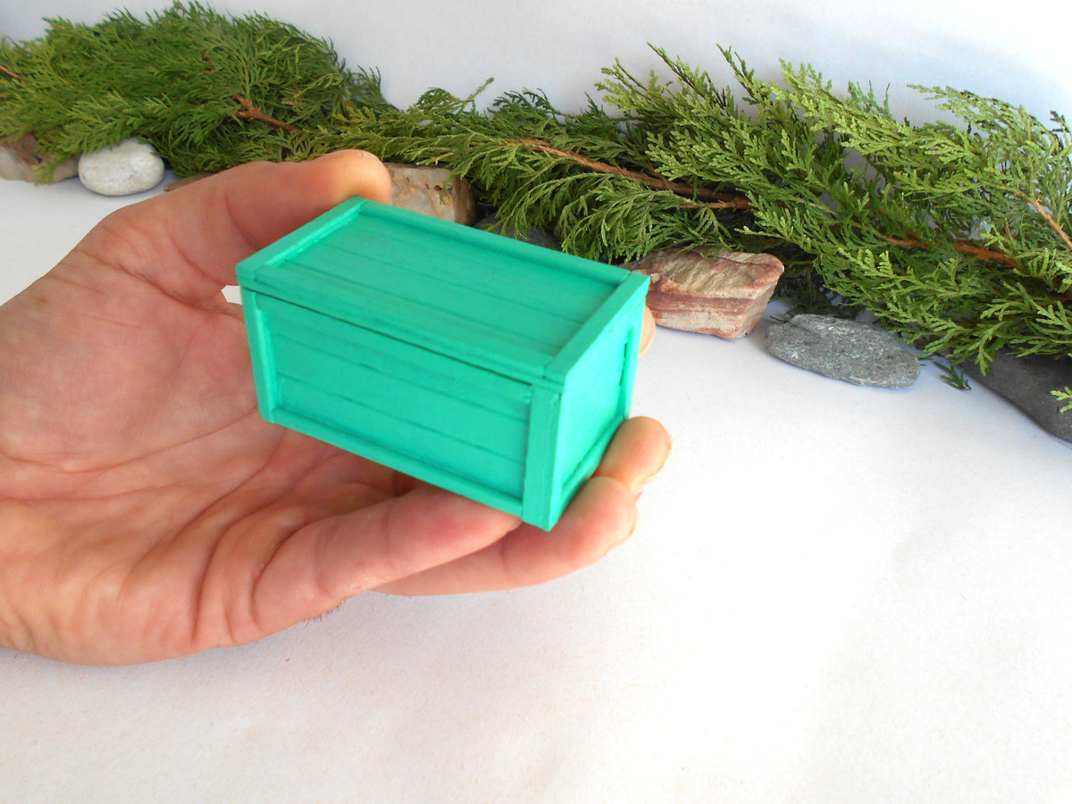A hand holding a small mint green wooden box with a lid, set against a white background with green pine branches and small stones.