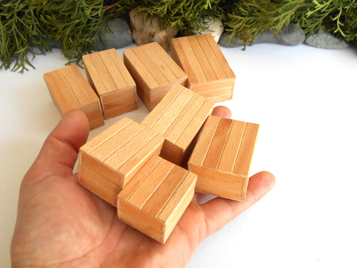A hand holding several small rectangular wooden boxes with grooved sides, with more blocks arranged behind on a white surface and greenery in the background.