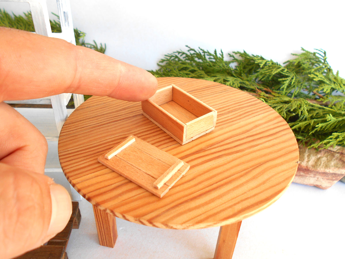A close-up of a miniature wooden table with a finger pointing at a small wooden box, with a matching wooden lid placed in front. The scene includes green foliage in the background.