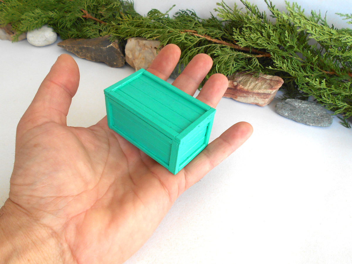 A miniature green wooden chest or box held in a person's hand, with greenery and small stones in the background.
