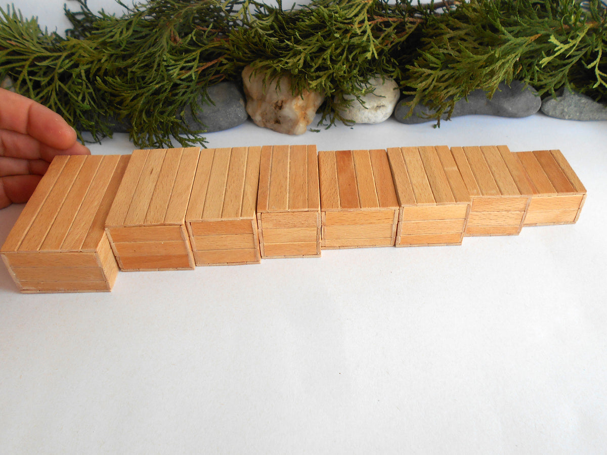 A set of natural light wood rectangular boxes arranged in a stepped row on a white surface, with a hand holding one block on the left and greenery and stones in the background.