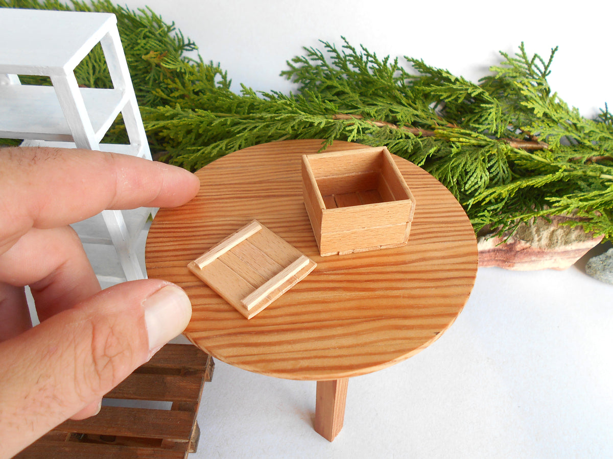 A miniature wooden box and matching tray displayed on a small round wooden table, with a human hand pointing at it for scale. The scene includes green foliage and a miniature white shelf in the background.