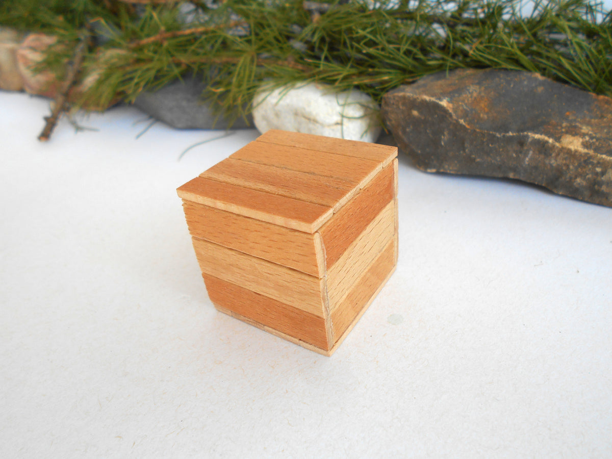 A small wooden cube-shaped box with a striped pattern of light and medium brown wood grain, placed on a white surface with pine branches and rocks in the background.