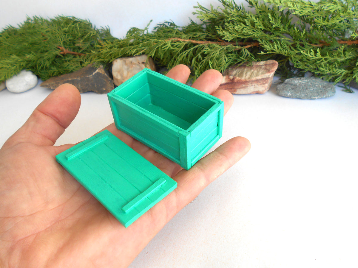 A hand holds a small turquoise green miniature storage box with its lid placed beside it. The background features a white surface with decorative green pine branches and small stones.