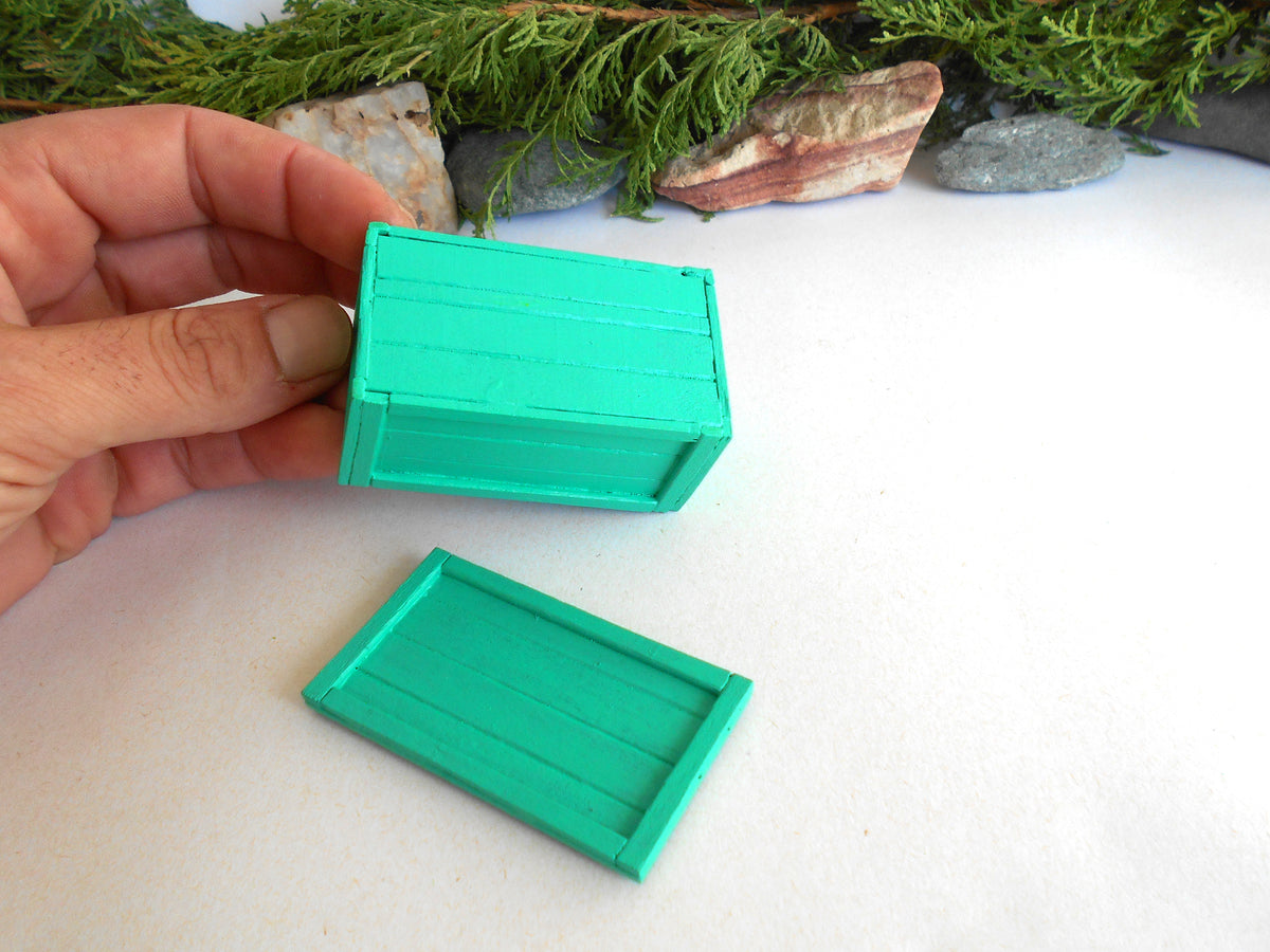 A hand holds a small painted green wooden box while a matching green lid lies on a white surface, with decorative stones and greenery in the background.