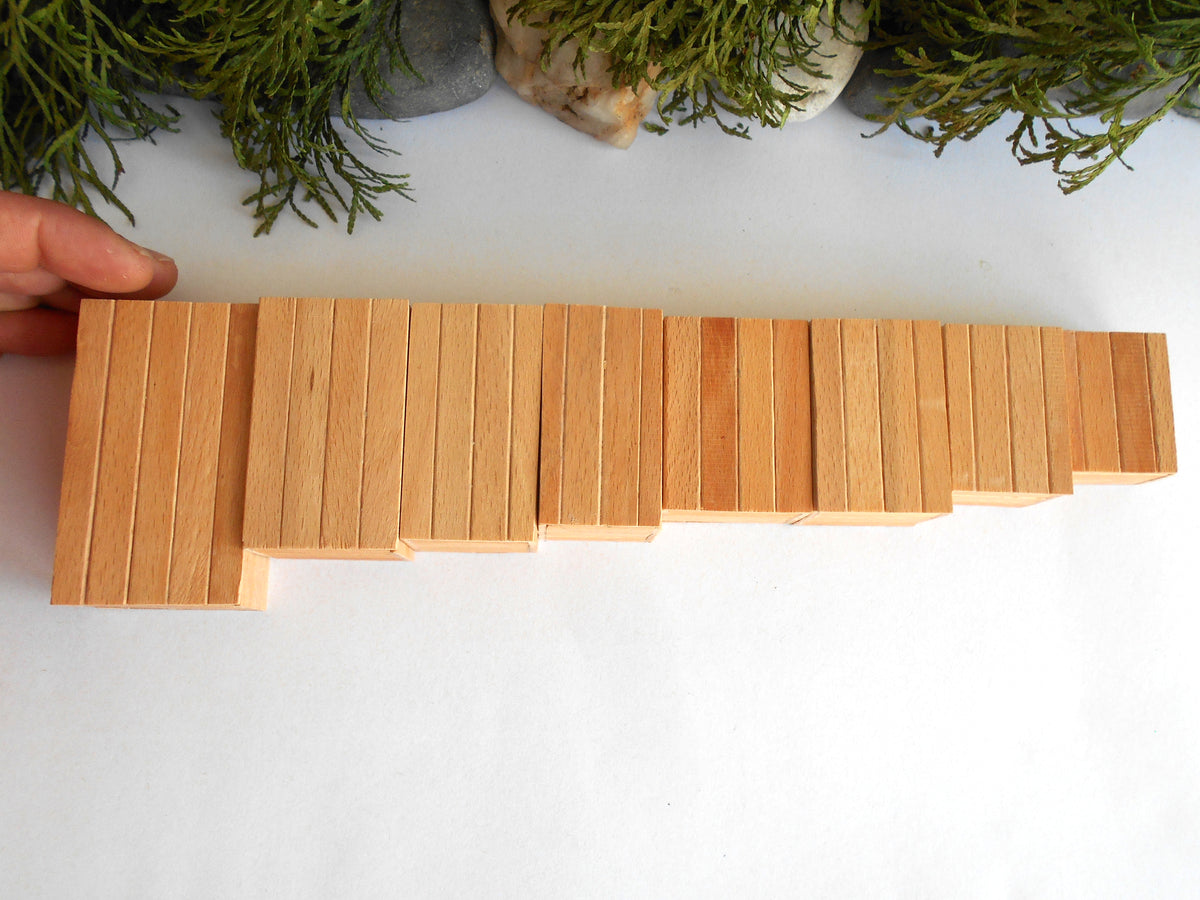A hand holds a long, rectangular wooden boxes toy made of many narrow light wood strips, photographed on a white surface with decorative greenery at the top.