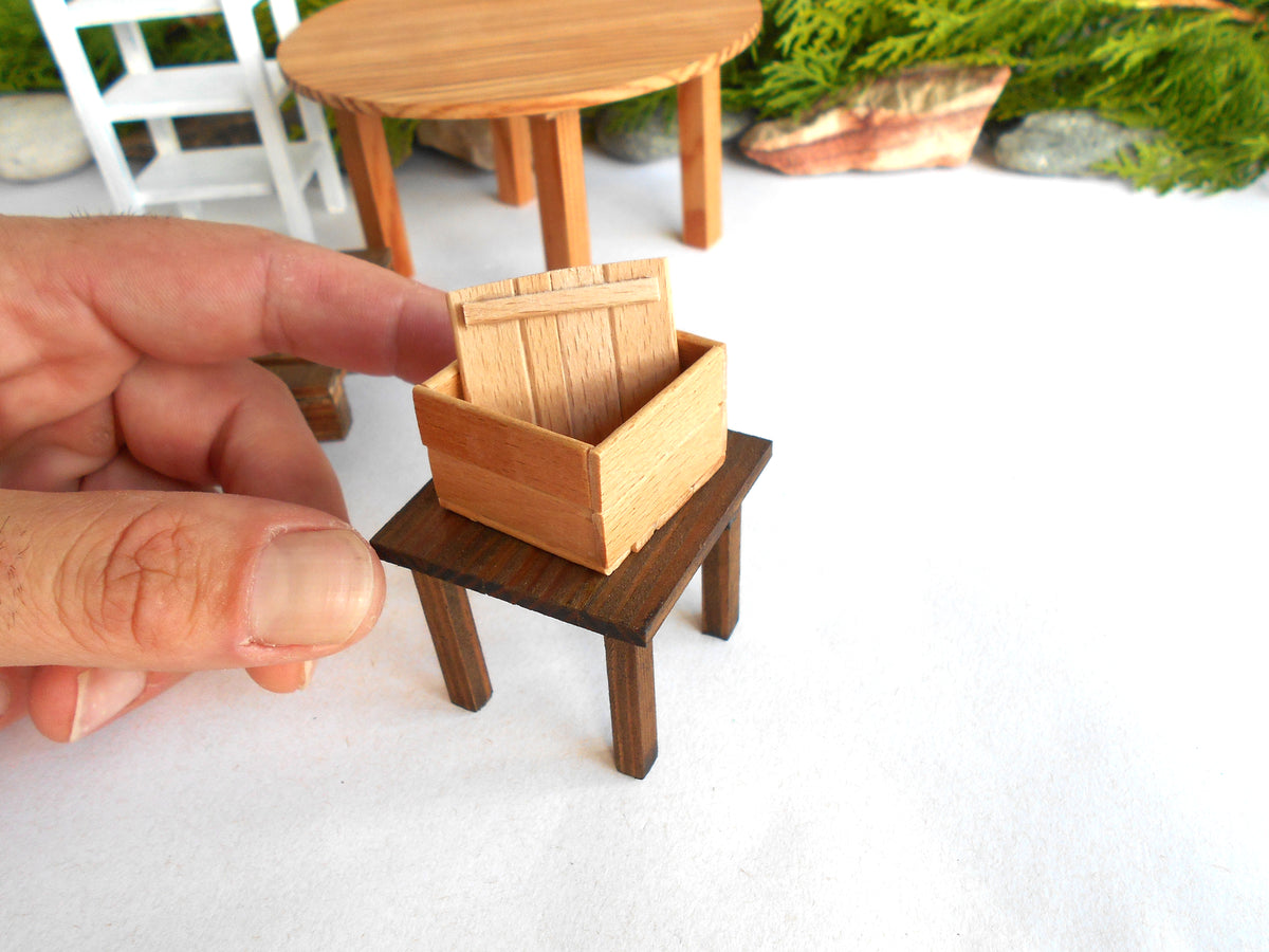 A miniature wooden crate model displayed on a small dark wooden stand, with a person's hand holding it for scale against a white background with miniature furniture props.