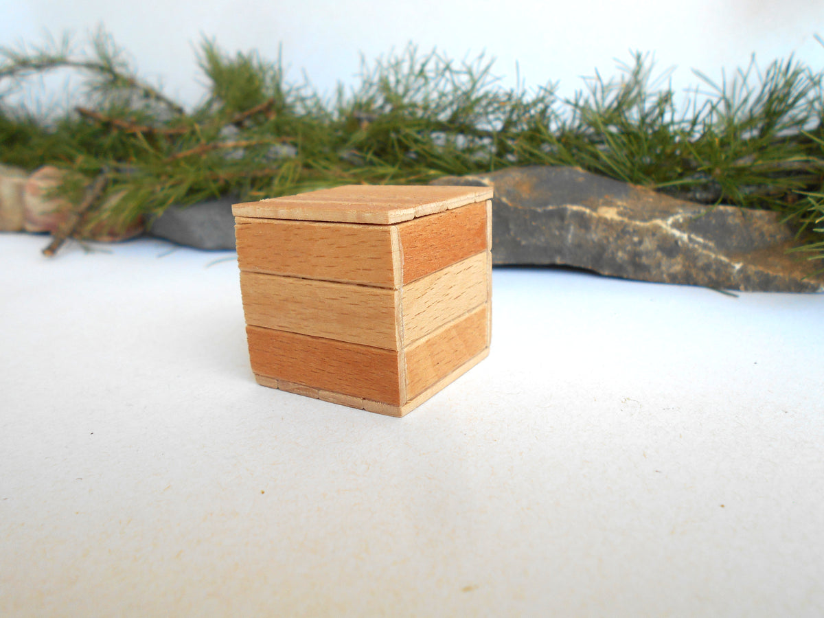 A small wooden box with a herringbone pattern made of alternating light and medium-toned wood strips, set against a white surface with blurred green pine branches and gray stones in the background.