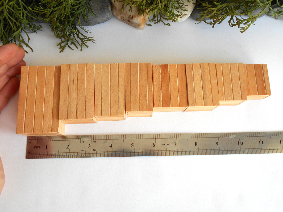 A set of light natural wood rectangular boxes arranged in a stepped row on a white surface, with a metal ruler underneath for scale and a hand holding the left side. Greenery and stones are visible at the top edge.