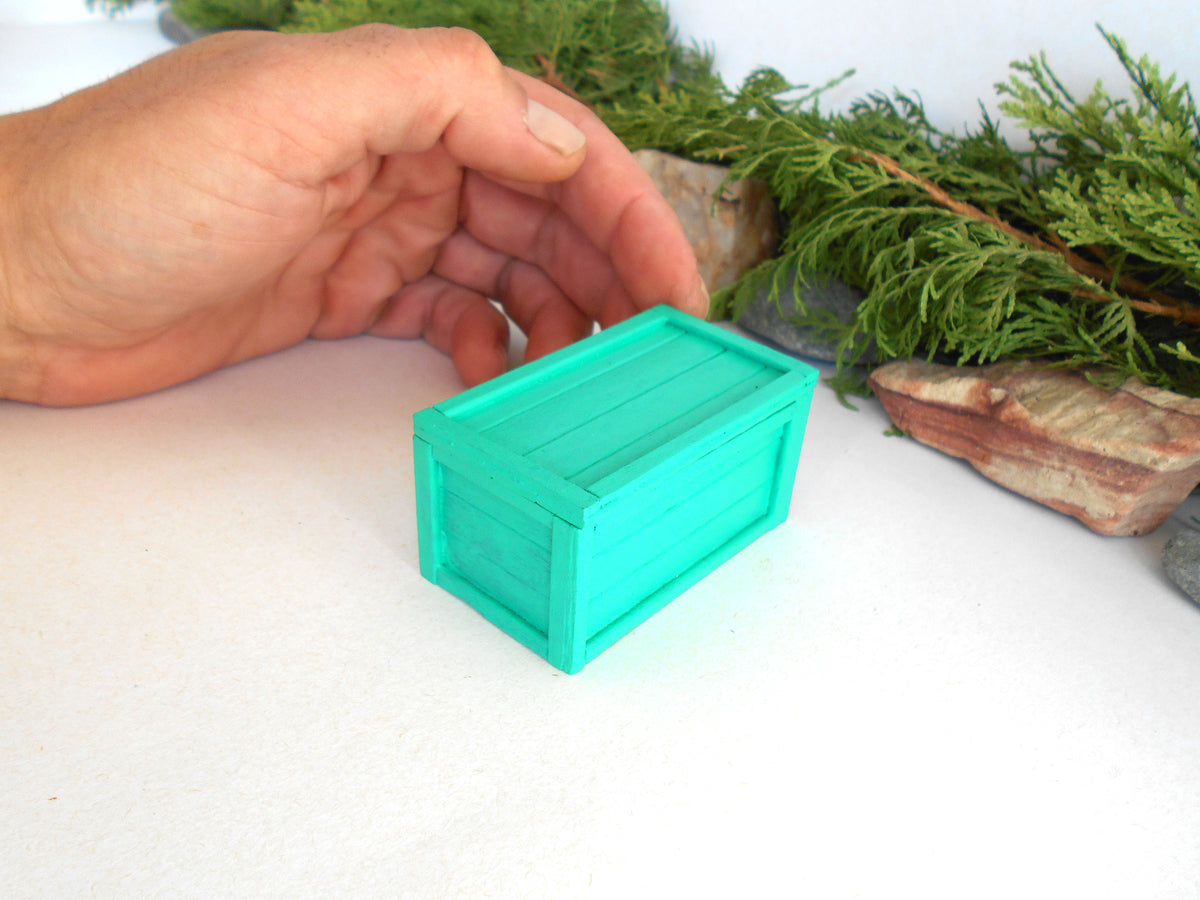 A hand holding a small turquoise wooden chest or box on a white surface, with green foliage and rocks in the background.