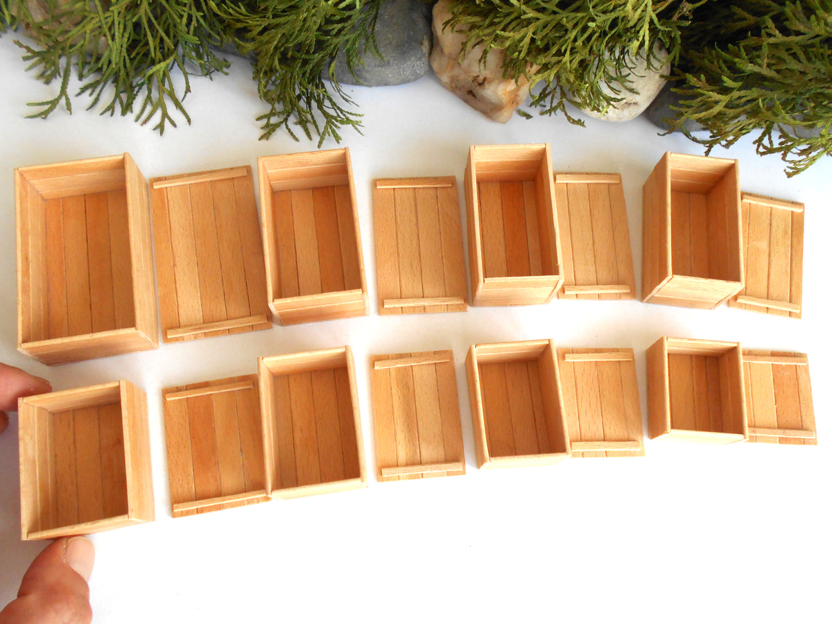A set of miniature light wood crate boxes arranged in two rows on a white surface, with green foliage and stones at the top and a hand holding one box at the bottom left.