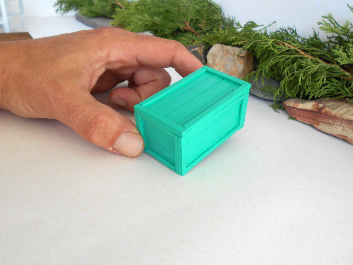 A hand holding a small mint green miniature wooden crate, set against a white tabletop with decorative green foliage in the background.
