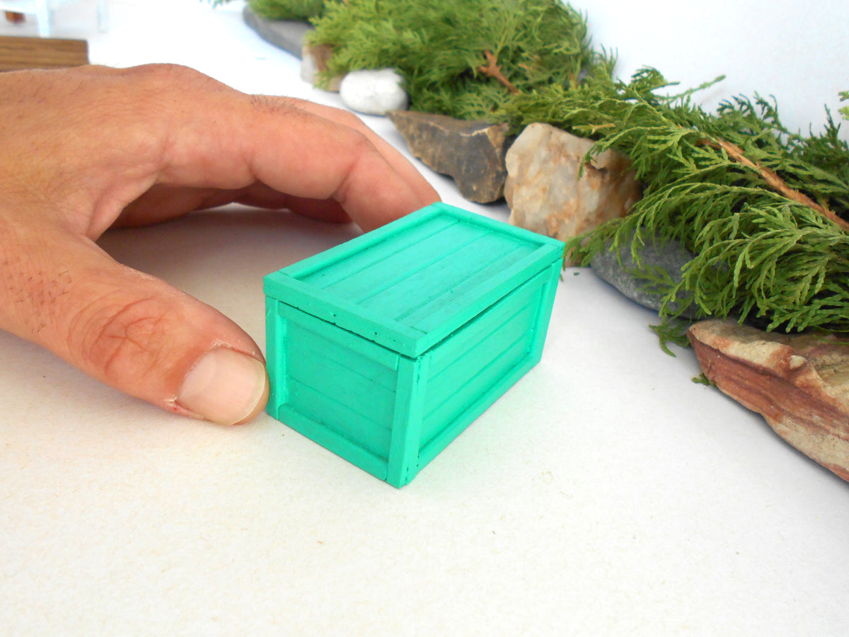 A miniature bright green plastic storage box being held by a person's hand on a white tabletop with miniature garden props in the background.