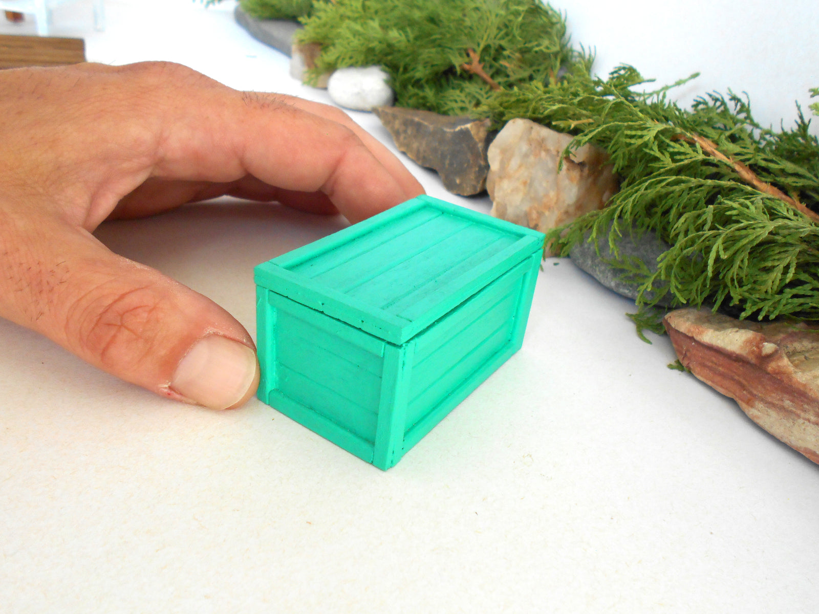 A miniature bright green plastic storage box being held by a person's hand on a white tabletop with miniature garden props in the background.