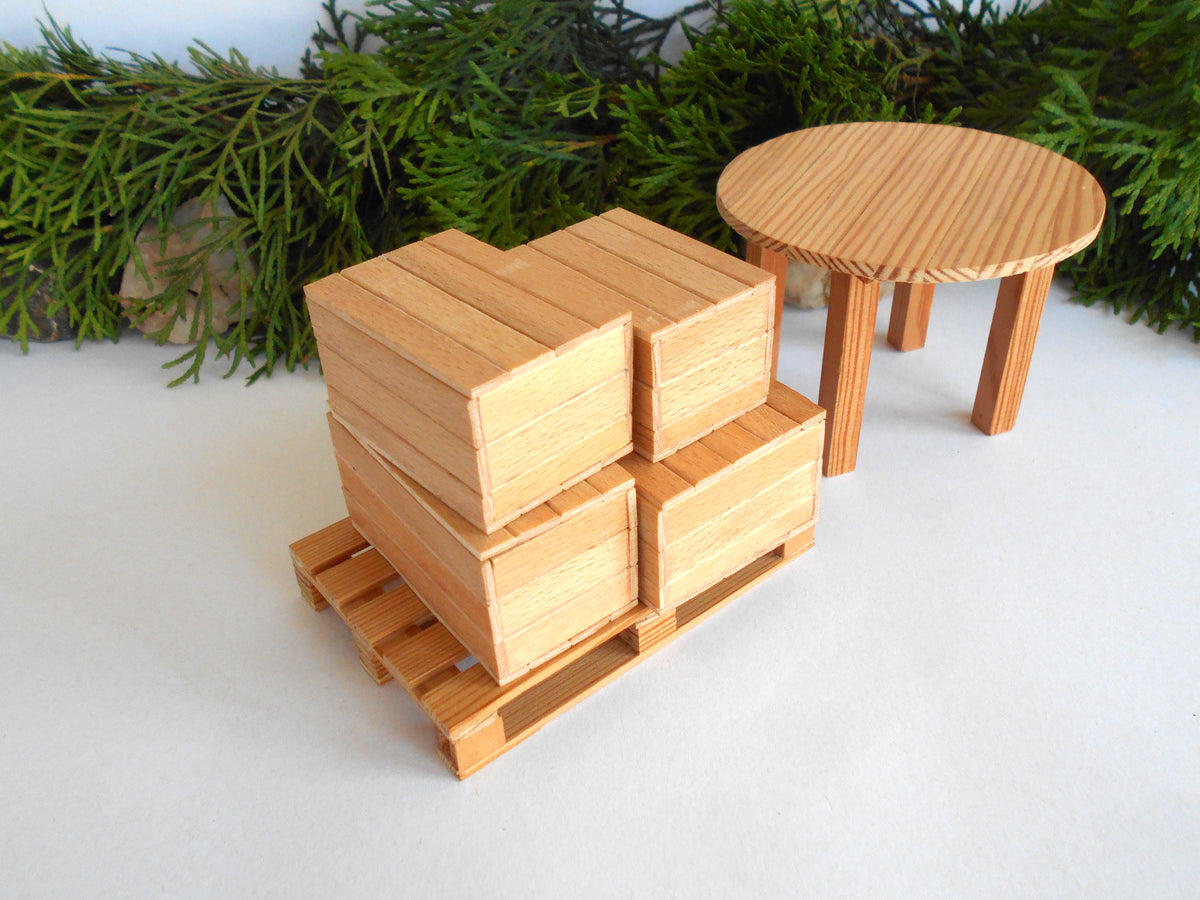 A miniature wooden toy set featuring a stack of four crates on a pallet and a small round table, displayed on a white surface with green pine branches in the background.
