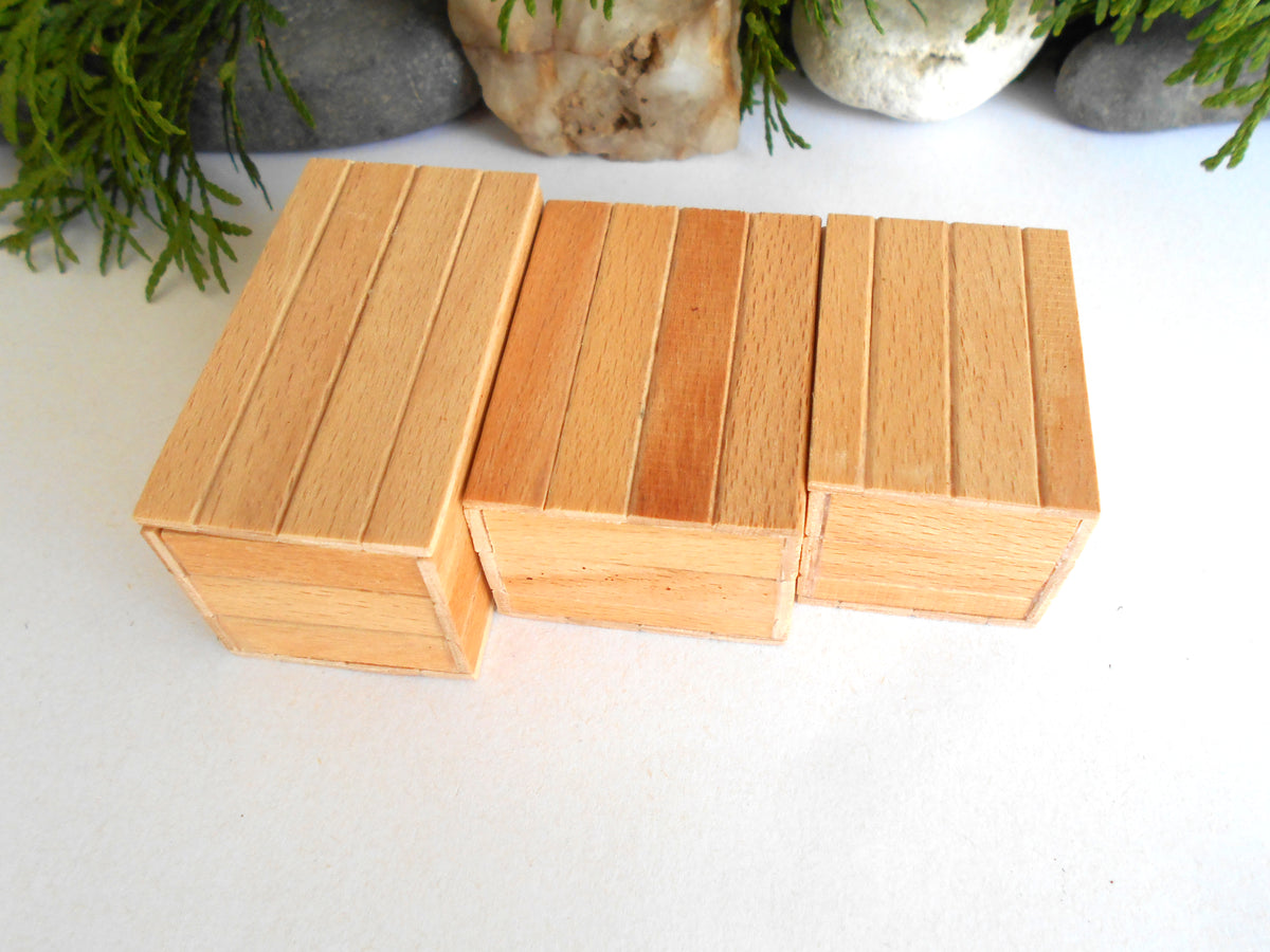 Three small rectangular wooden boxes with a slatted top design, arranged side-by-side on a white surface with decorative stones and greenery in the background.