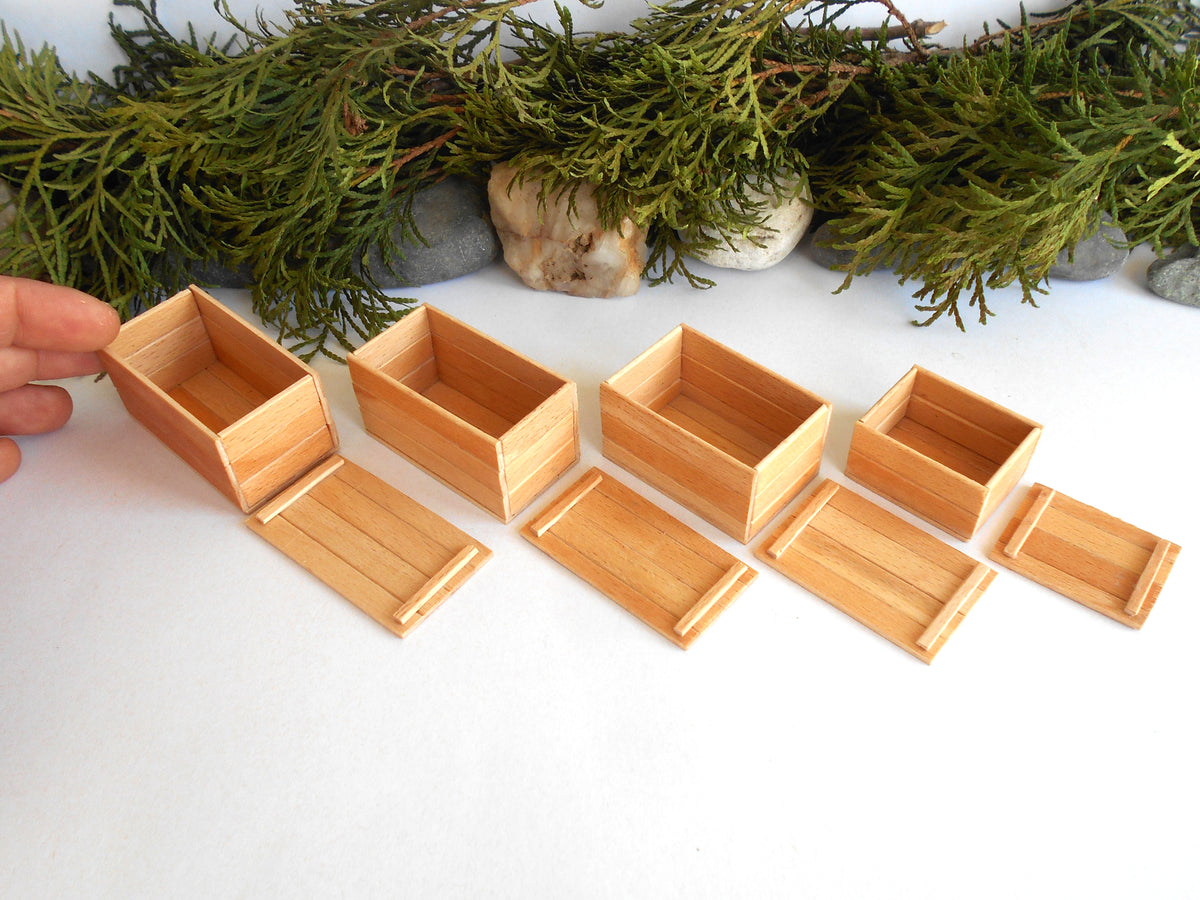 Six miniature light brown wooden boxes and trays with matching lids, displayed on a white surface with green foliage and small stones in the background. A hand is visible on the left holding one of the boxes.