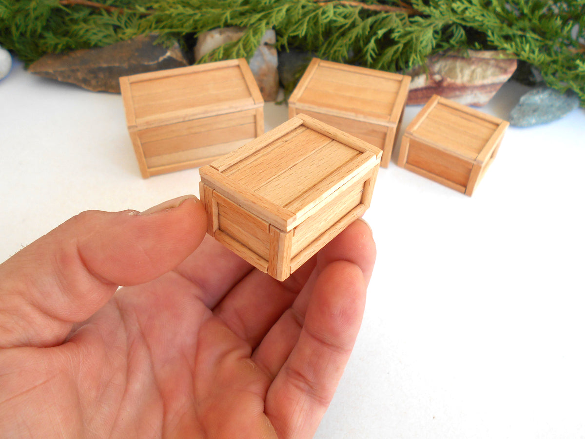 A hand holding a miniature light wood crate box, with four similar small wooden storage boxes arranged in the background on a white surface with green foliage and stones.