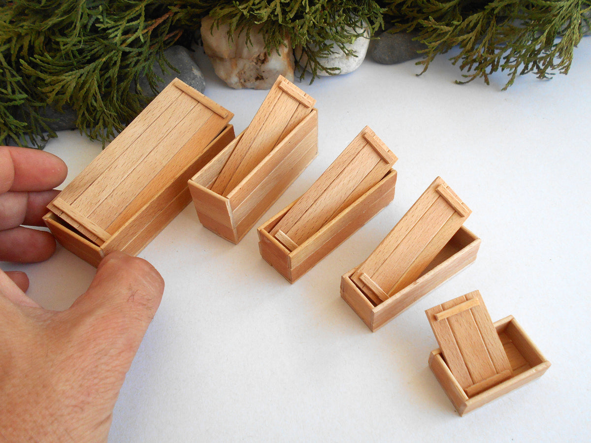 Five miniature light wood coffin-shaped boxes of different sizes arranged on a white surface, with a hand holding one of the larger boxes and evergreen branches in the background.