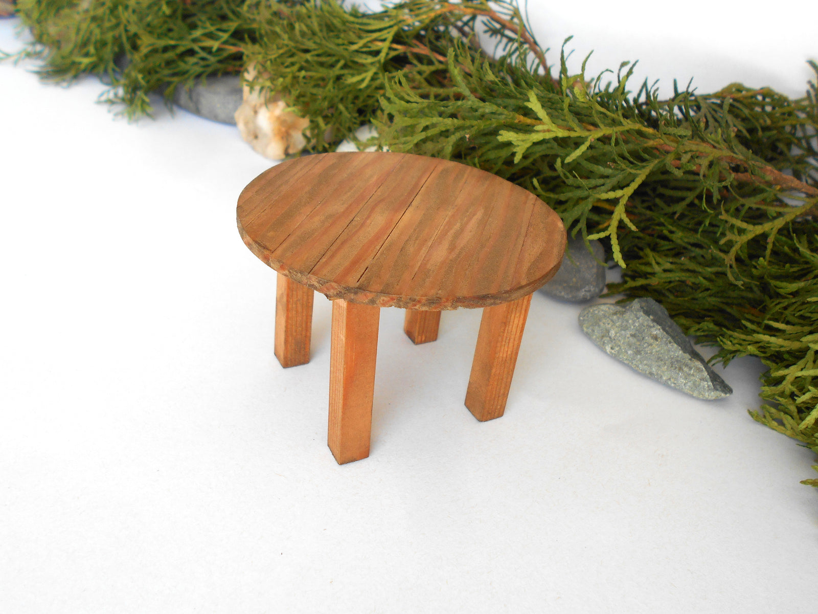 A miniature round wooden stool or table with four legs, styled with green foliage and small stones on a white background.