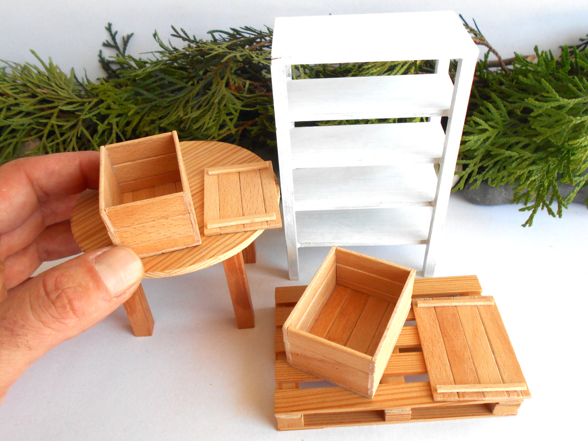 A miniature dollhouse-scale wooden crate set with a round table, a white shelving unit, and pallet-style bases, shown with a hand holding one crate on a white surface with greenery in the background.