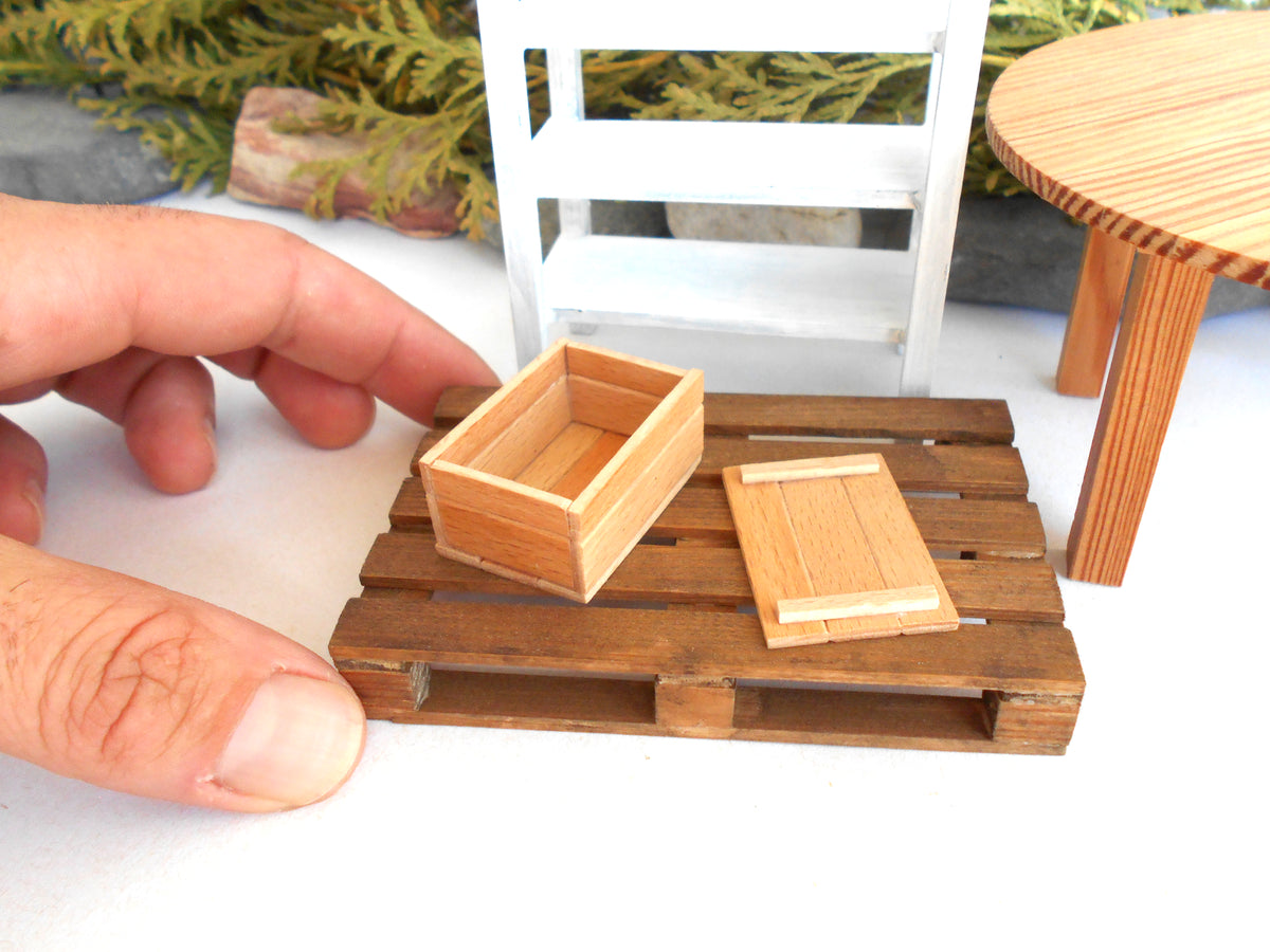 A close-up of a miniature wooden crate and matching lid set on a tiny wooden pallet, with a human hand holding the pallet for scale. The scene includes miniature furniture in the background, such as a white shelf and a round wooden table.