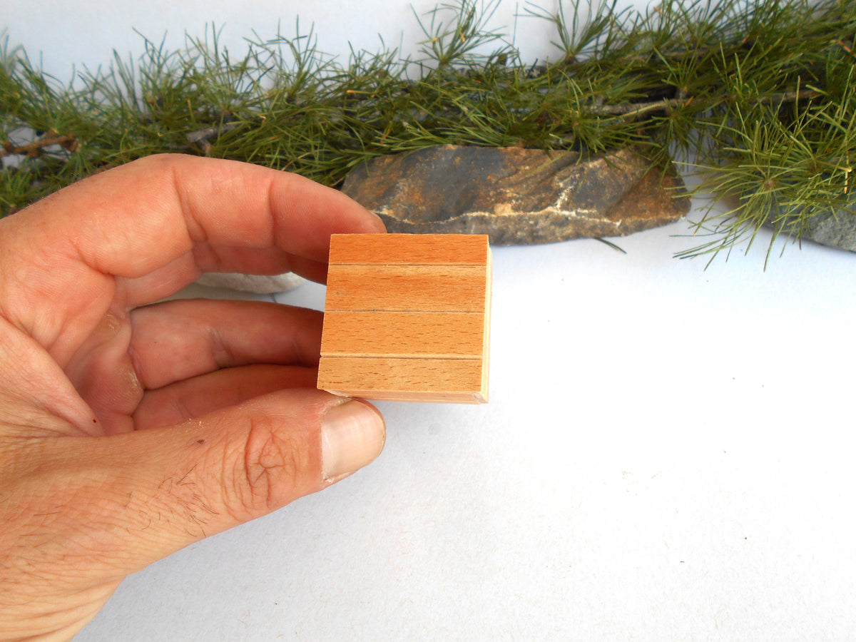 A hand holding a small rectangular wooden block with three horizontal grooves, set against a white background with pine branches and a stone prop.